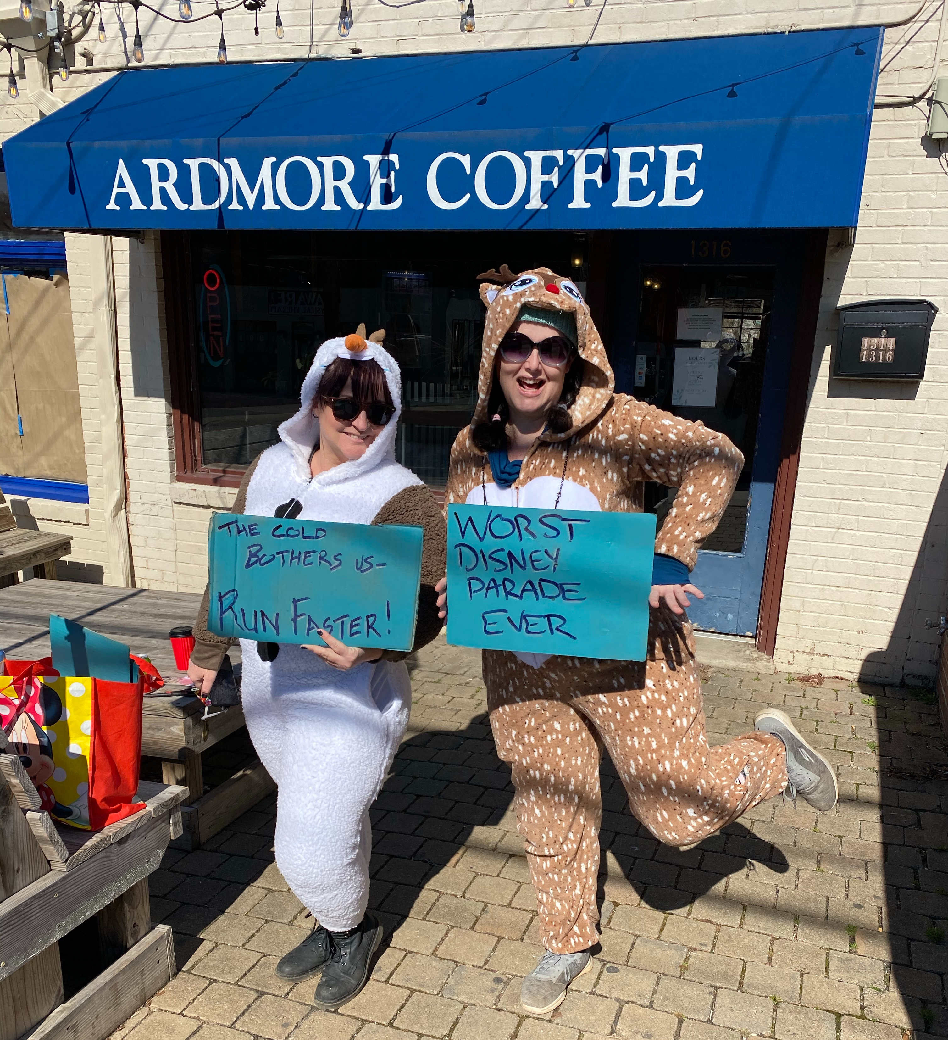 Women in Olaf and reindeer onesies outside a coffee shop. Olaf holds a sign saying "the cold bothers us, run faster", the deer's sign says "worst Disney parade ever".