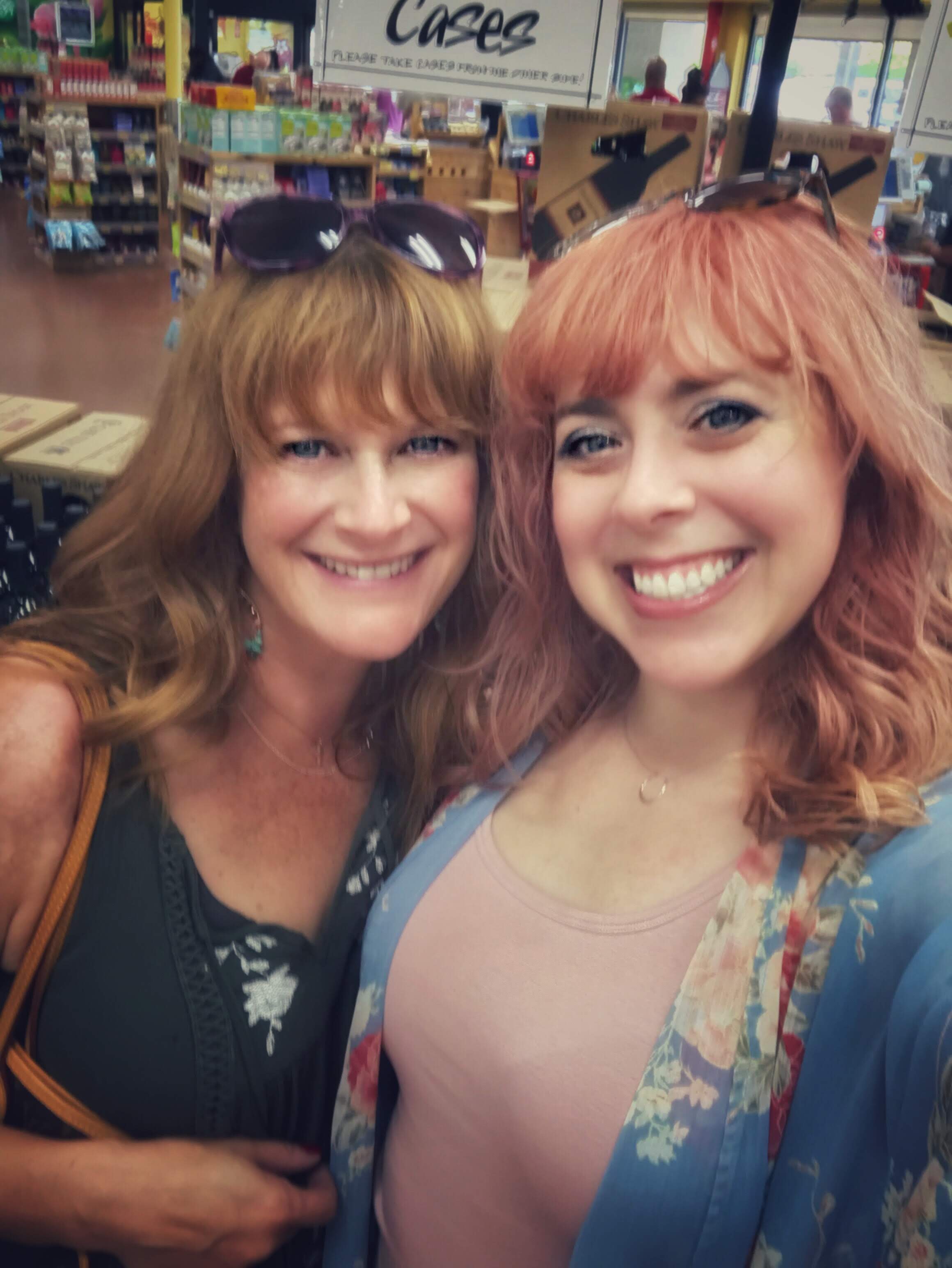 two women in the wine section of a Trader Joe's. Both have wavy hair and bangs, with sunglasses on top of their heads. The one on the left has auburn hair and a green floral top. The one on the right has rose gold hair with a pink tank top and blue floral kimono.