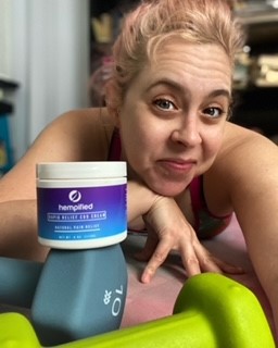 Woman lying on her stomach on a yoga mat. Her light hair is pulled up and messy. Product balanced on a light blue 10 lb. weight. A smaller bright green weight is in the foreground.